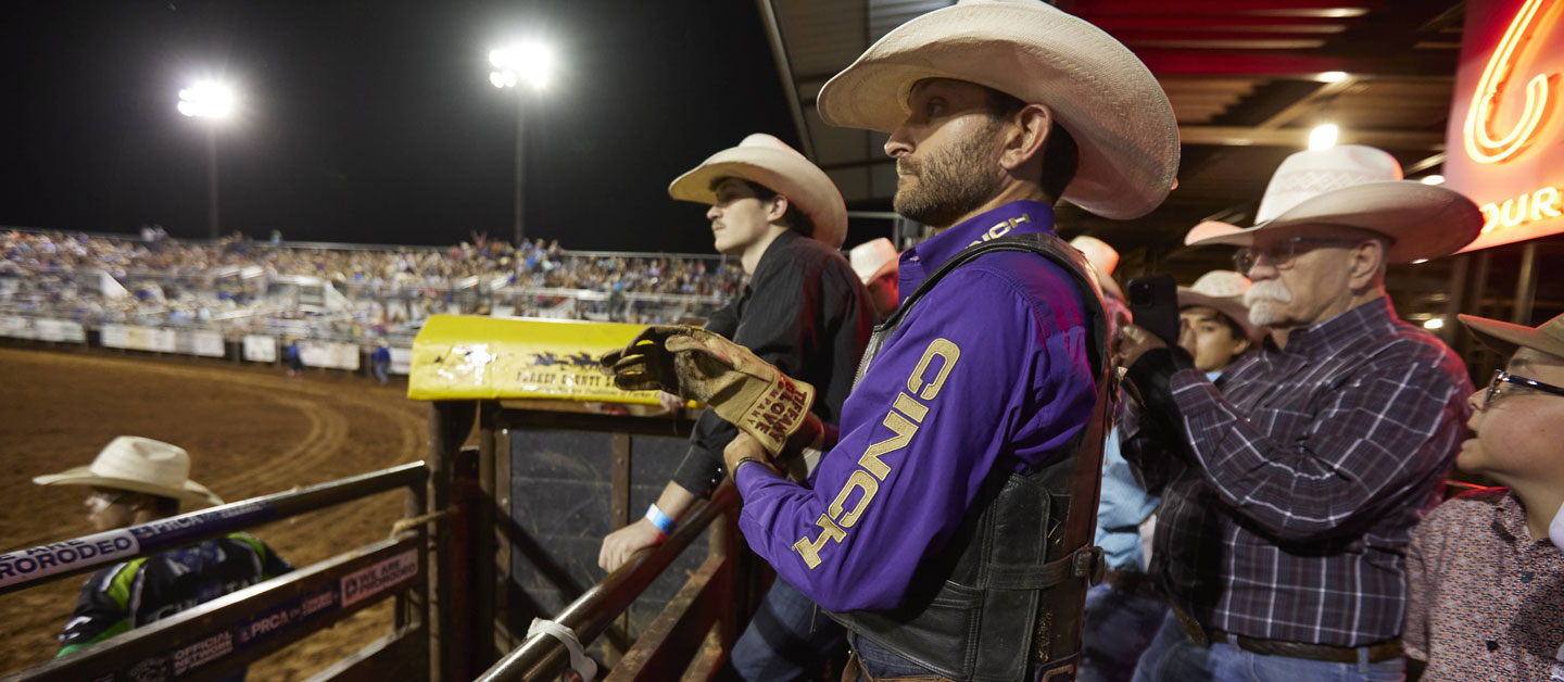 Man wearing a purple shirt and cowboy hat standing on the bucking chutes at a rodeo.